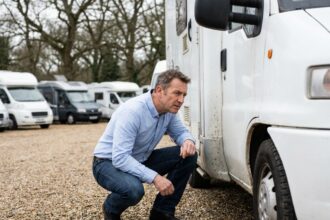 Homme inspectant un camping-car d'occasion blanc dans un parc de revendeurs, vérification des joints extérieurs lors d'un déstockage